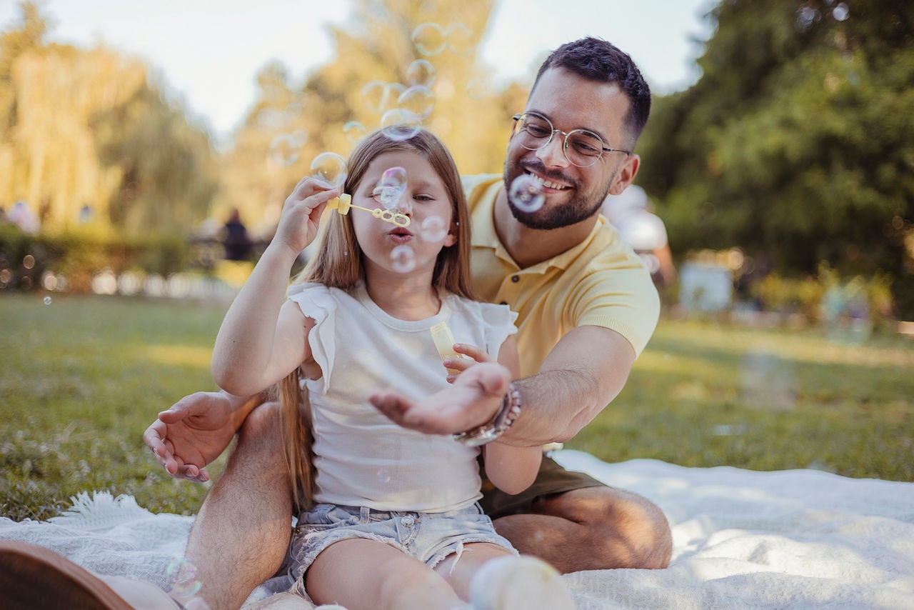 Outdoor Portrait of Little Daughter With Her Father Making Bubbles in the Public Garden at Vacation Day or Holiday. Family Concept.