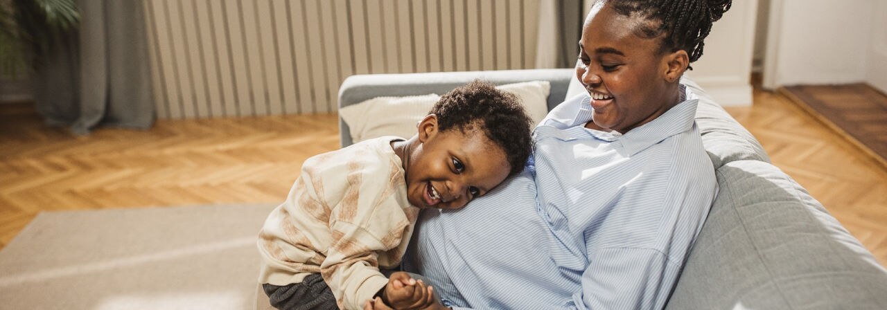 Cute little boy touching belly of his pregnant mom while relaxing together on sofa in living room.