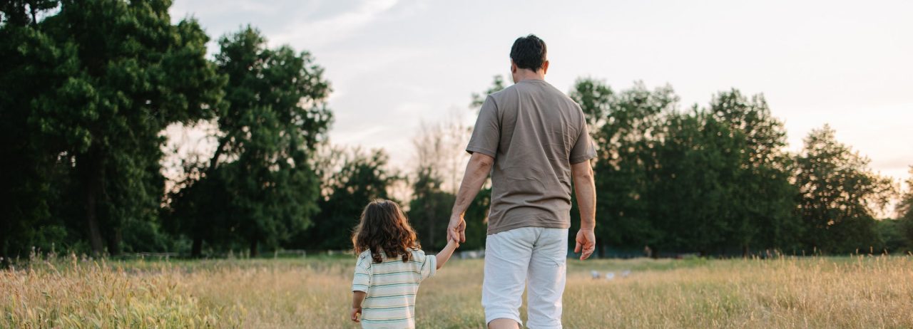 A young dad and his son are walking in the grass field while holding hands.