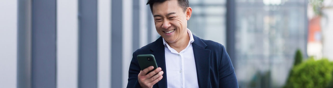 asian male freelancer walking near business center holding phone, smiling reading news, successful businessman
