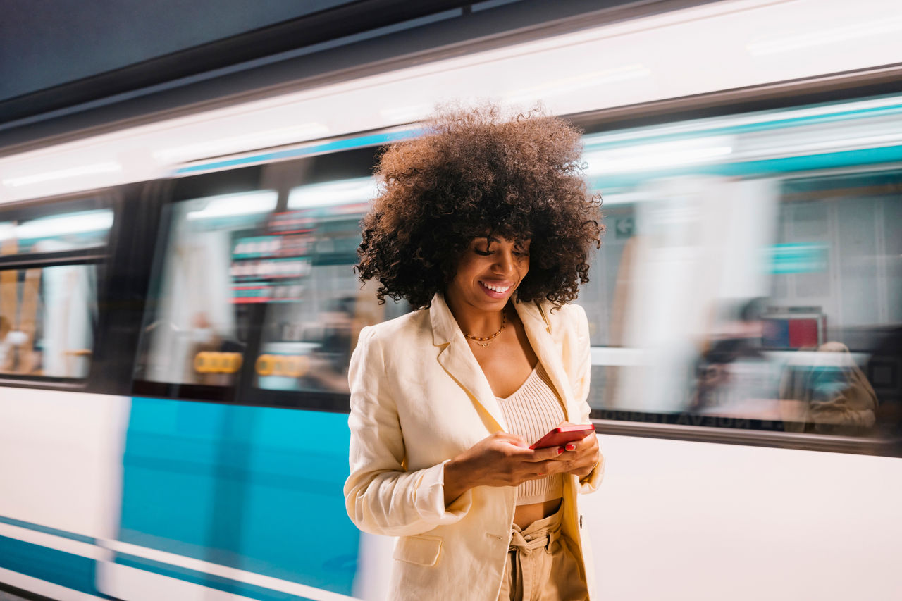 Smiling woman using smart phone in front of moving subway train; Shutterstock ID 2191420627; purchase_order: P&B Loop 1&2(26)-1*.16.01.2024; job: ; client: ; other: 