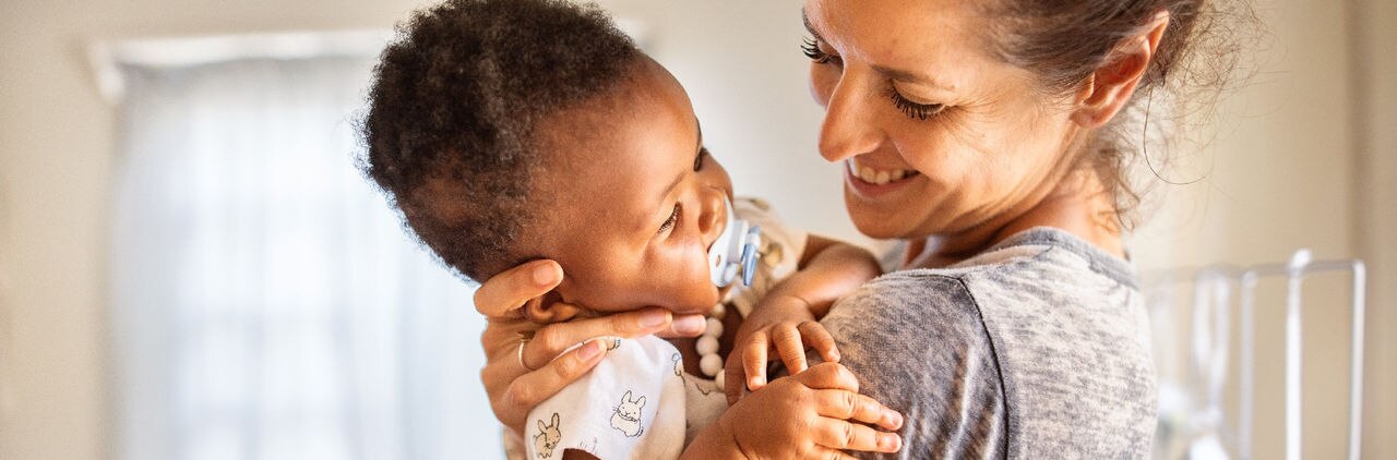 Smiling mom holding her adorable little adopted baby girl with a pacifier in her mouth in her arms at home