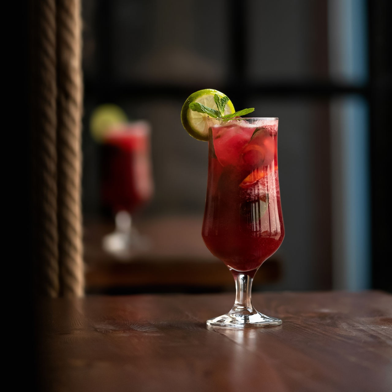 Glass of red Alcoholic cocktail drink with mint leaf and lime or lemon slice on dark blurred background. Side view. Copy space.