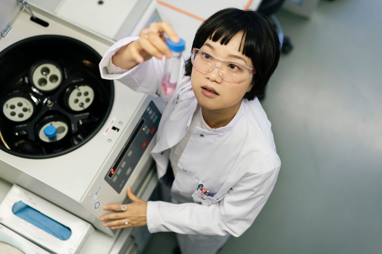 A asian female scientist is working inside a laboratory. She's taking a close look and examining a flask that cotains a pinkish liquid.