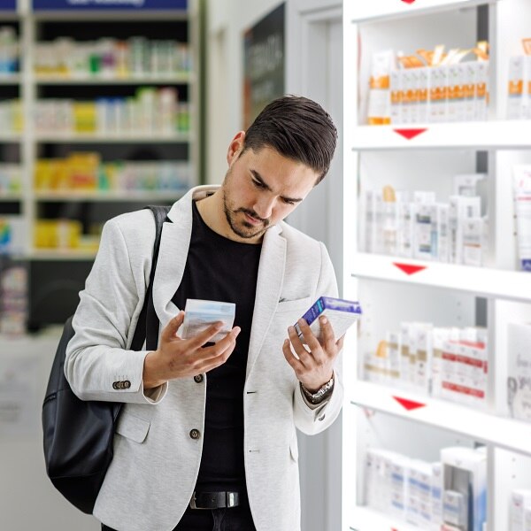 A young Caucasian businessman is standing in a pharmacy and choosing betweeen two boxes of pills.