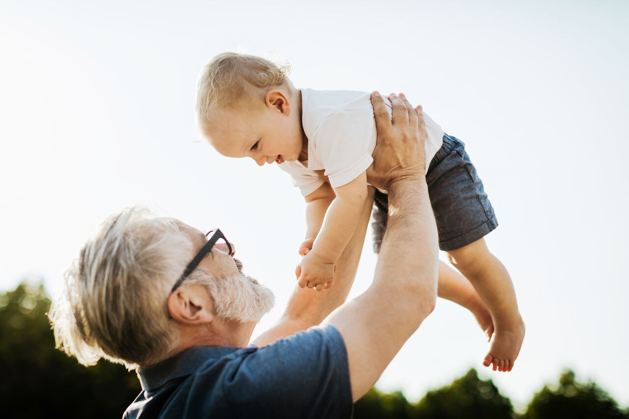 A grandpa holding his young grandchild in the air while on a day out together at the park.
