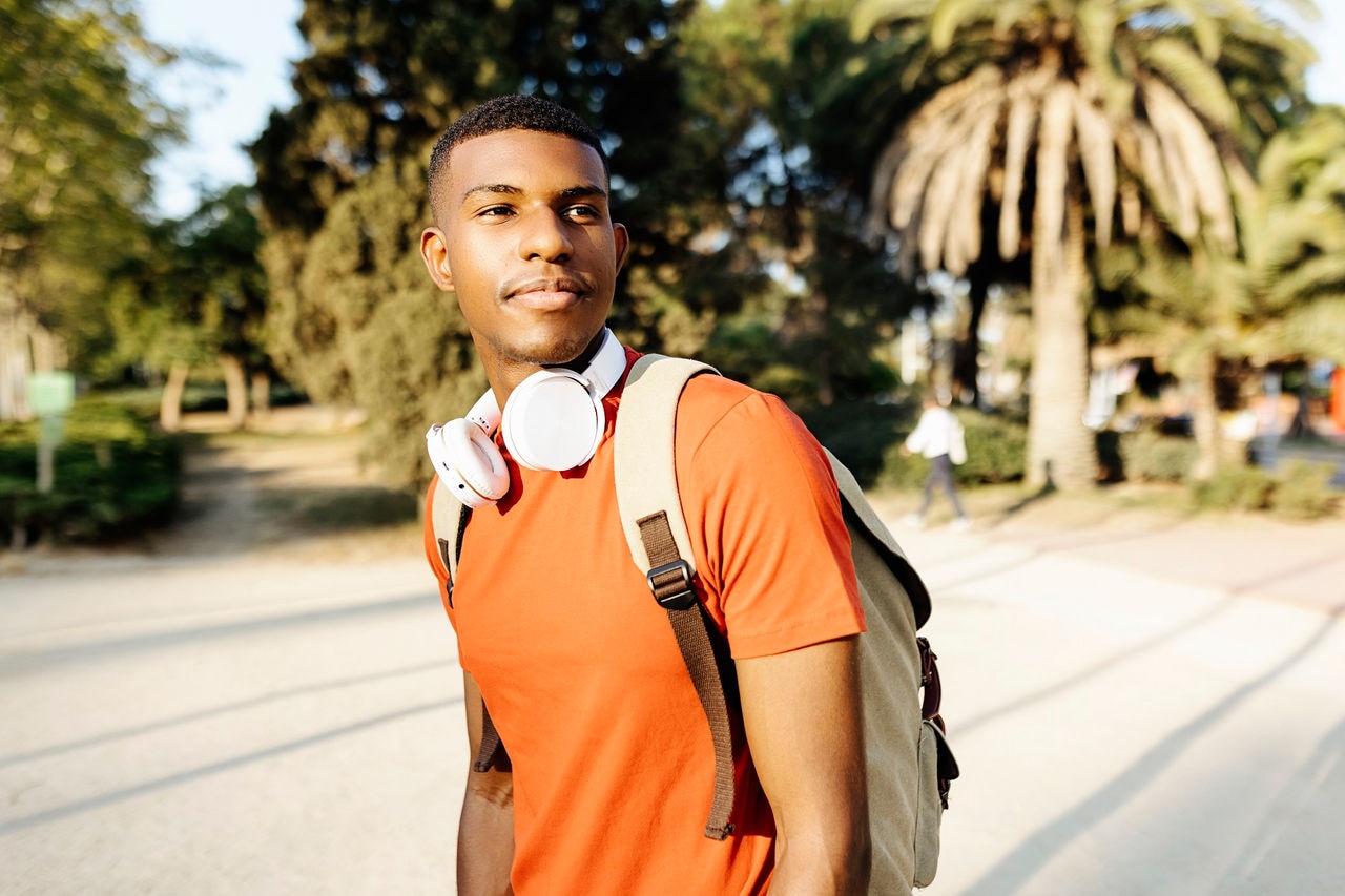 Cropped shot of a young black man student with headphones and bag walking in public park