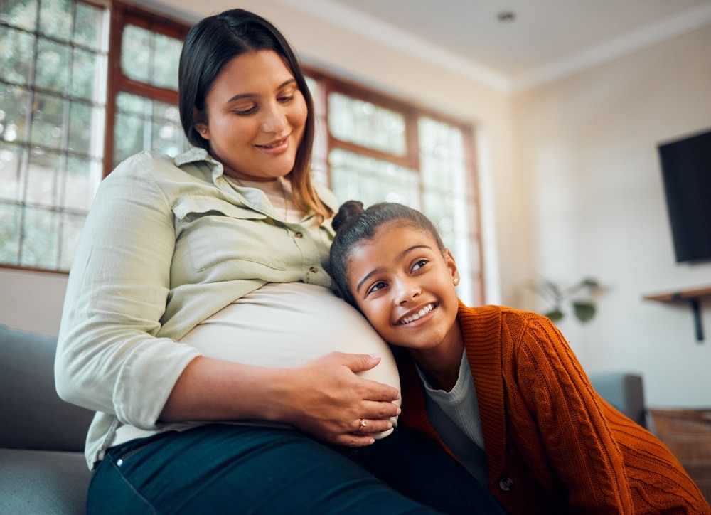 Love, home and pregnant mother with child listening to movement of baby in belly with happy smile. Family home, pregnancy and mom relaxing with excited, young and curious daughter on sofa
