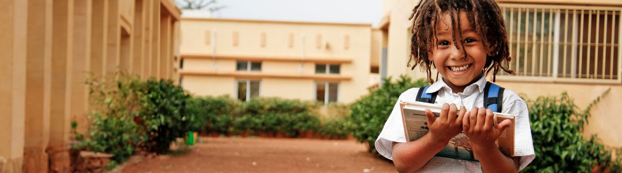A school boy wearing dread locks is standing in the school yard, holding books in his two hands, facing the camera and smiling.