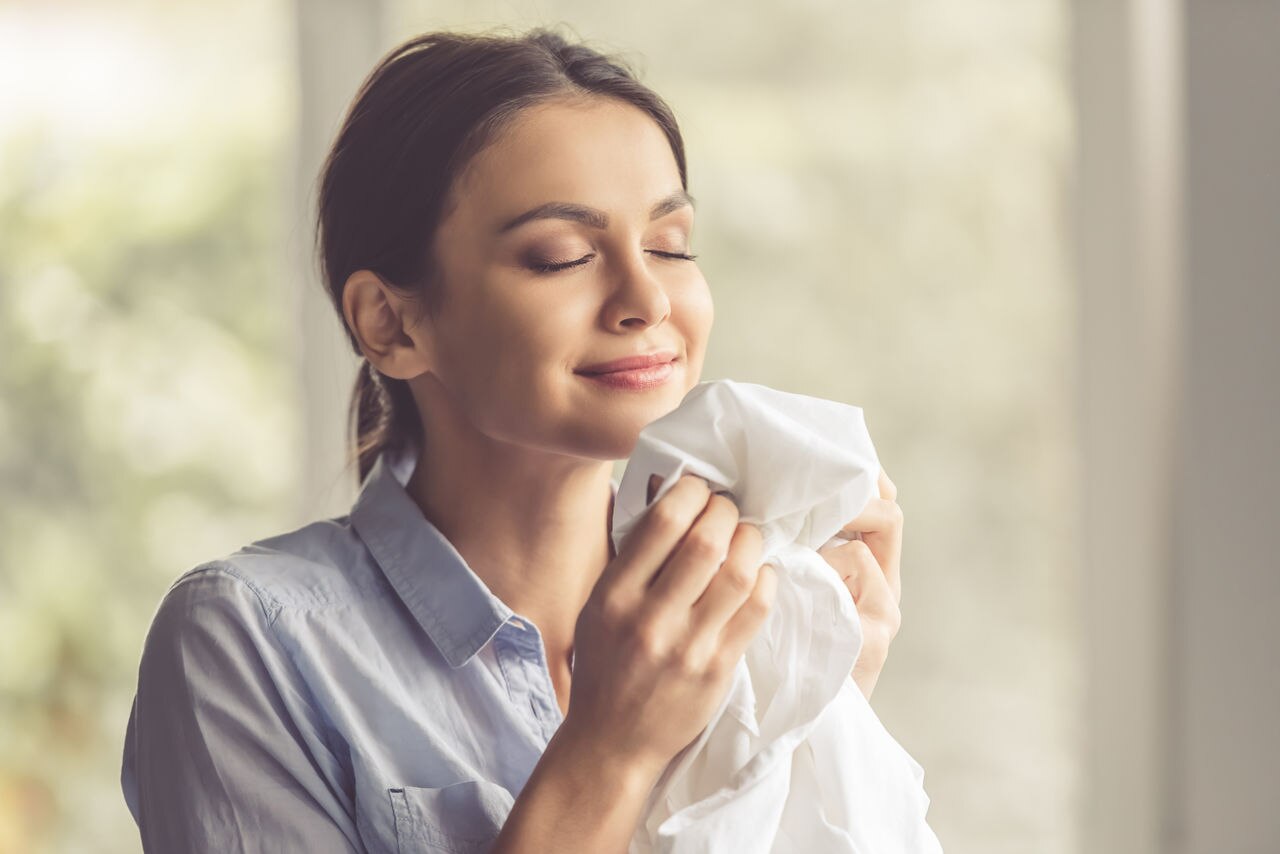 Beautiful young woman is smelling clean clothes and smiling while doing laundry at home