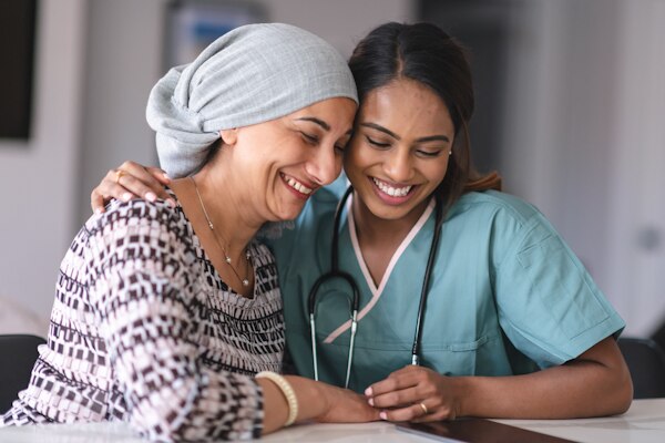 Portrait of an Indian woman fighting cancer with her physician. The doctor is a mixed-race female. The two women are seated next to each other indoors. They are embracing and laughing.