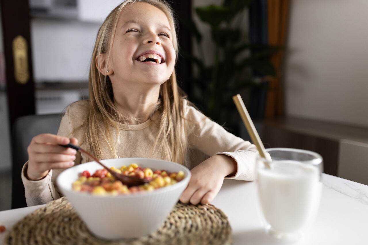 Cute little caucasian girl sitting at table on kitchen early morning and preparing breakfast with colorful cornflakes and milk. Kid enjoying life with healthful food, healthy lifestyle concept